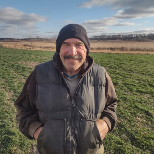 smiling man wearing winter clothing with his hands in his jacket pockets standing on the edge of a rye field with blue sky and a couple of clouds in the background