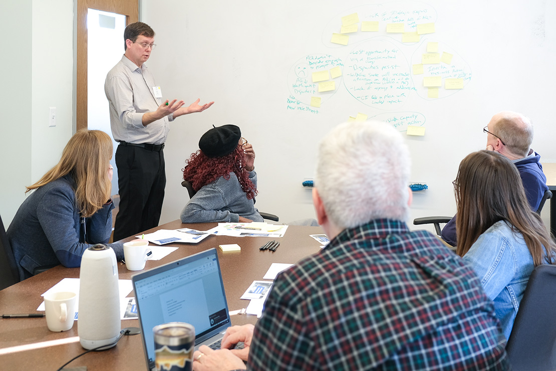 A group of six colleagues gathered around a conference table in a bright meeting room. At the head of the table, a man in a light striped shirt and glasses stands gesturing toward a whiteboard covered in clusters of yellow sticky notes and hand‑drawn diagrams. Seated around the table are four other adults—two women and two men—listening attentively; one woman rests her chin on her hand, another types on a laptop. On the table are printed handouts, pens, coffee mugs, and a tall speaker. The scene conveys an engaged brainstorming session.
