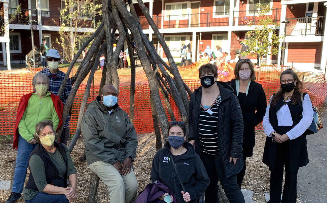 A group of eight people standing in front of a teepee made of natural branches