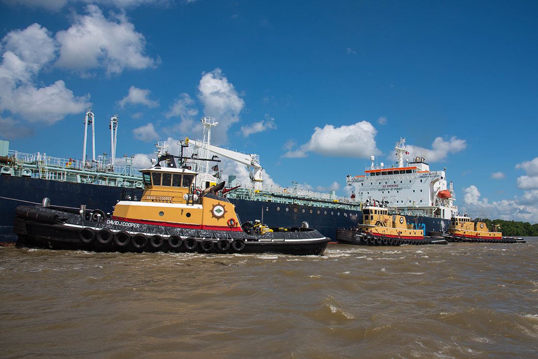 Three yellow and black tugboats providing tug escort to large, blue marine vessel on the Mississippi River near New Orleans.