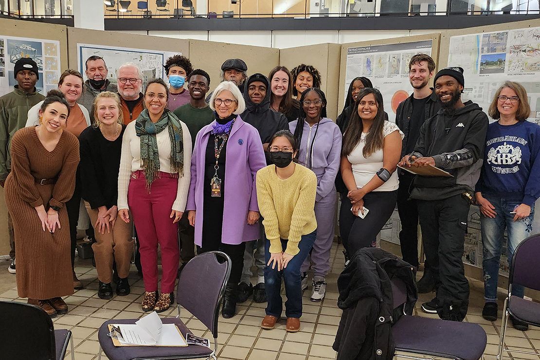 A group of high school students, North Minneapolis community members, and University of Minnesota School of Architecture students pose for a photo in a bright room. A wall with design project posters is behind the group.