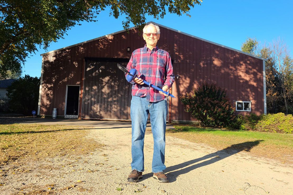 Chuck Louis stands in front a red barn while holding a soil probe tool. He is wearing a red flannel shirt and jeans.