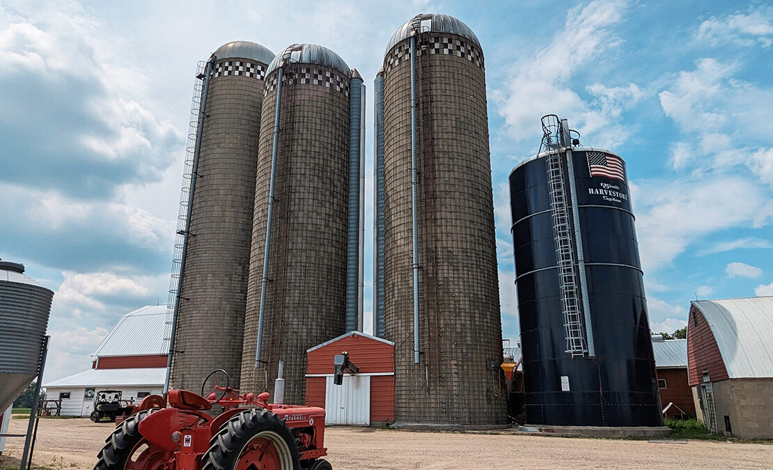 Four tall grain silos are seen on a farm. A red tractor is parked in front of the silos.