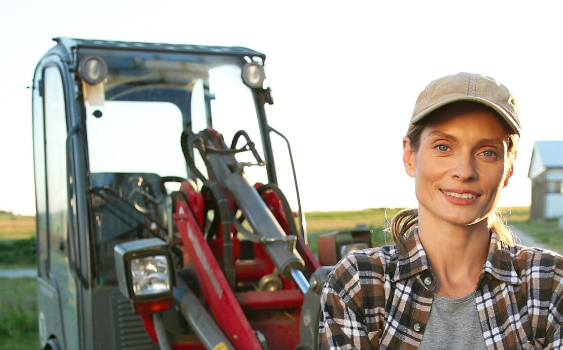 White female farmer wearing a plaid shirt and hat standing in front of a red tractor next to a farm field.