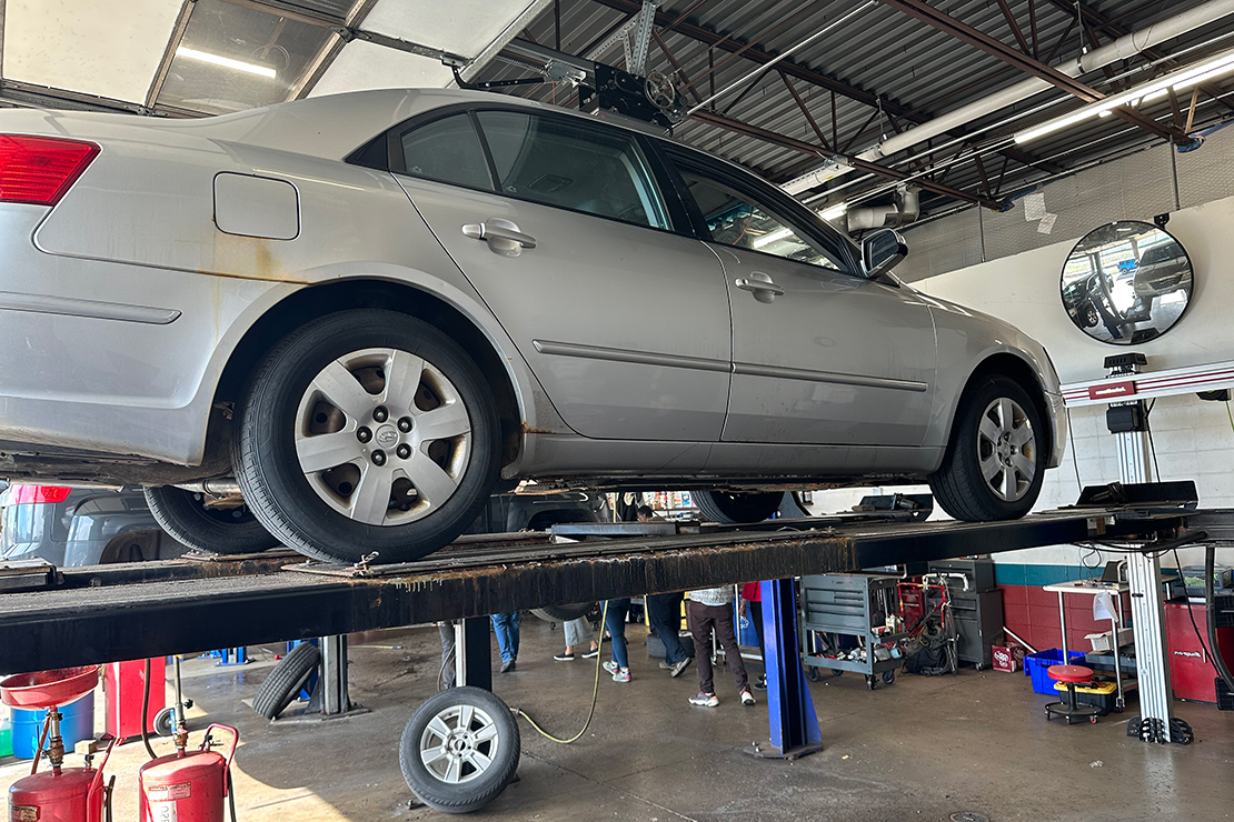 A silver car is elevated on a vehicle lift inside an auto repair shop. The shop is equipped with various tools, equipment, and a large mirror mounted on the wall. Several people can be seen in the background working.