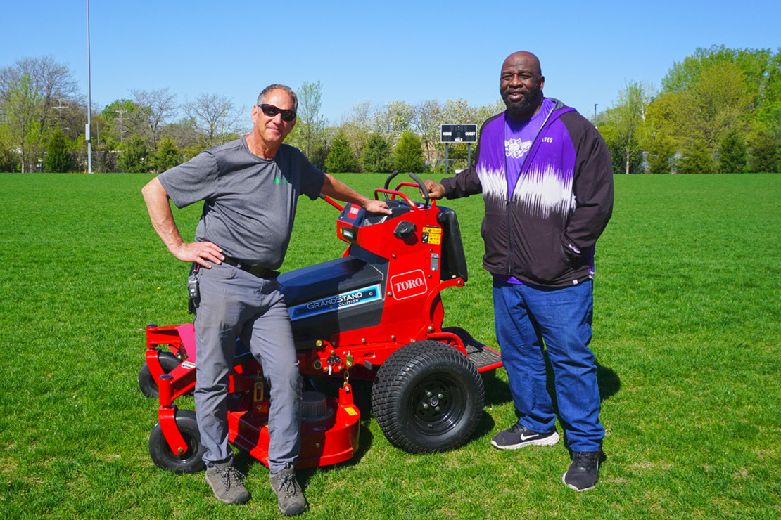 Greg Kroth, Parkway Lawn Service and Dwyane Waltower, Hiawatha Academies standing on a soccer field next to a commercial electric lawnmower. Greg is wearing sunglasses, a gray shirt, and gray pants. Dwyane is wearing a black and purple jacket and blue jeans.