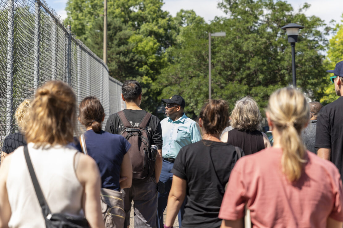 A group of people walks outdoors, led by a man in a turquoise shirt and black cap, next to a chain-link fence.