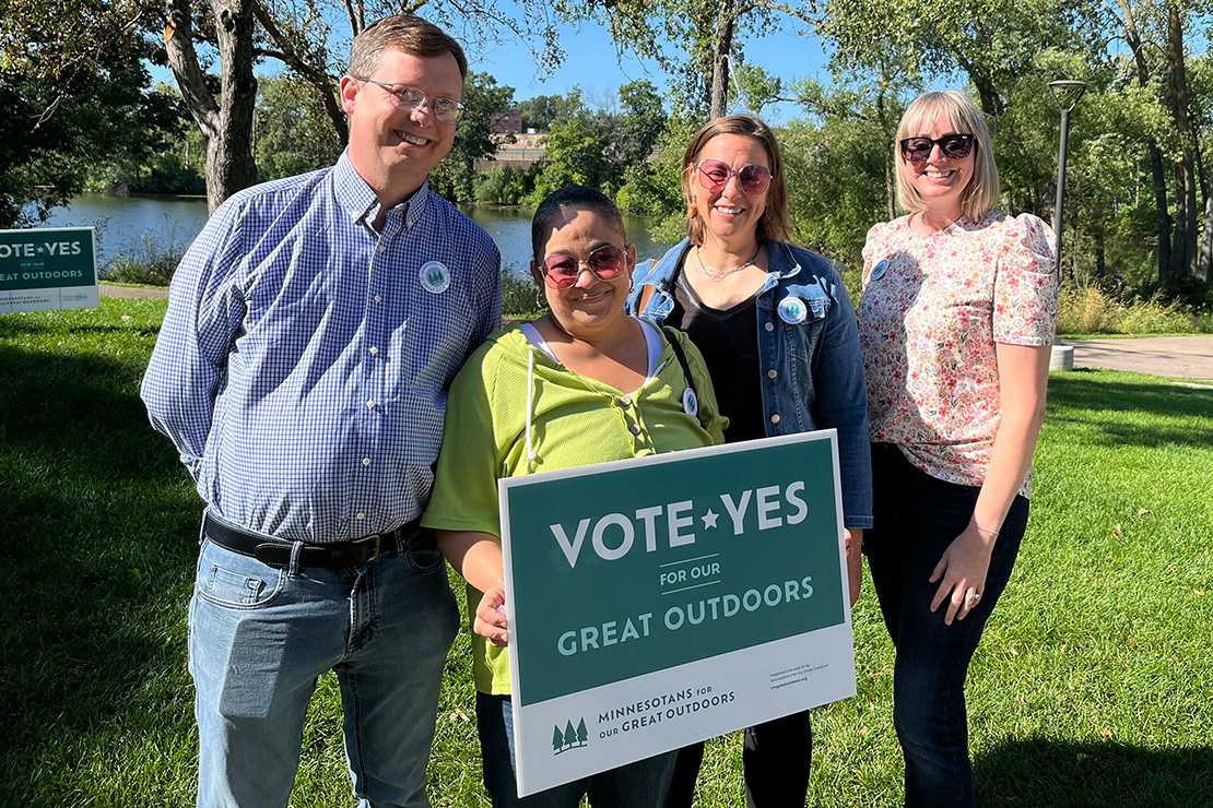 Four individuals smiling and standing together outdoors on a sunny day, holding a sign that reads 'Vote Yes for Our Great Outdoors.' They are supporting the Minnesota ballot measure to renew lottery proceeds for environmental conservation. Trees and a lake can be seen in the background.