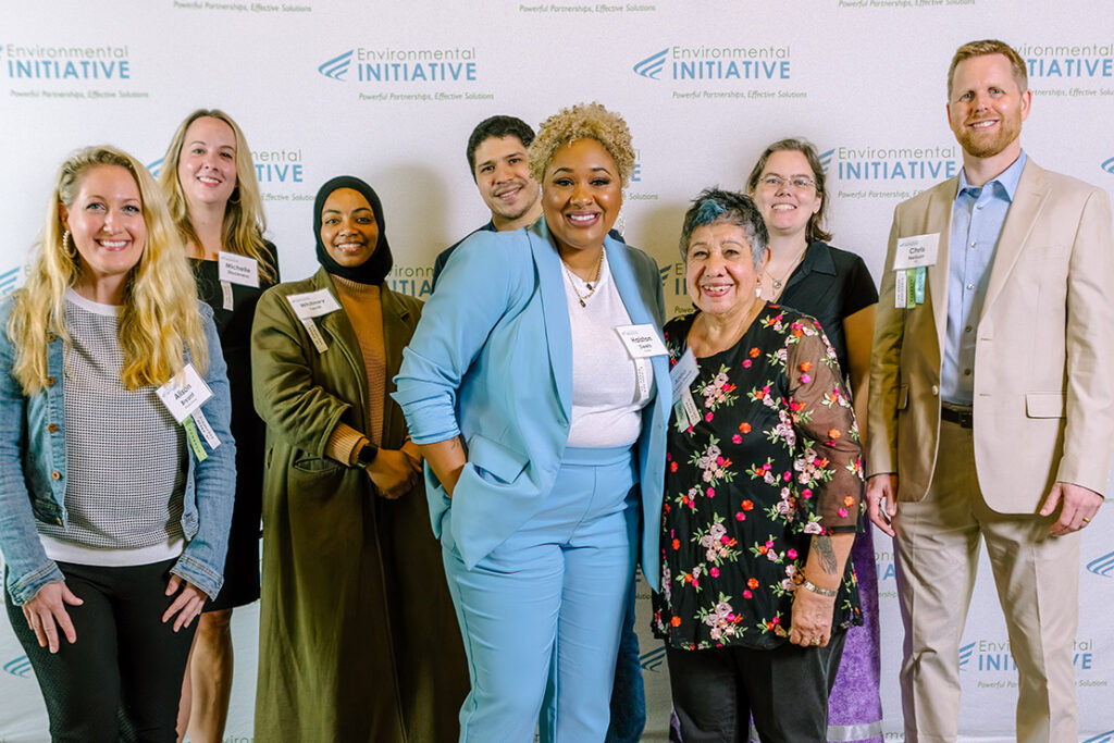 A group of eight people, standing and smiling in front of a backdrop with the Environmental Initiative logo. They are dressed in a variety of styles, including business and casual attire, and represent a diverse range of backgrounds and ages.