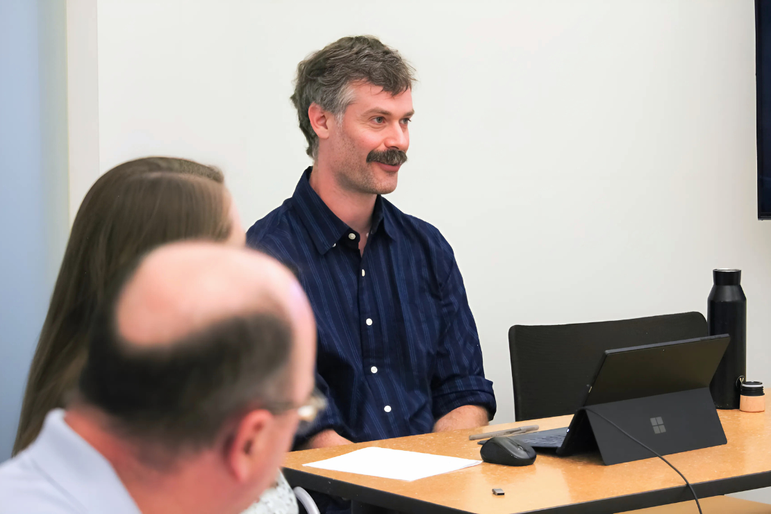 A man with a mustache sits at a table with a tablet and water bottle, engaged in a discussion with others in a meeting room.