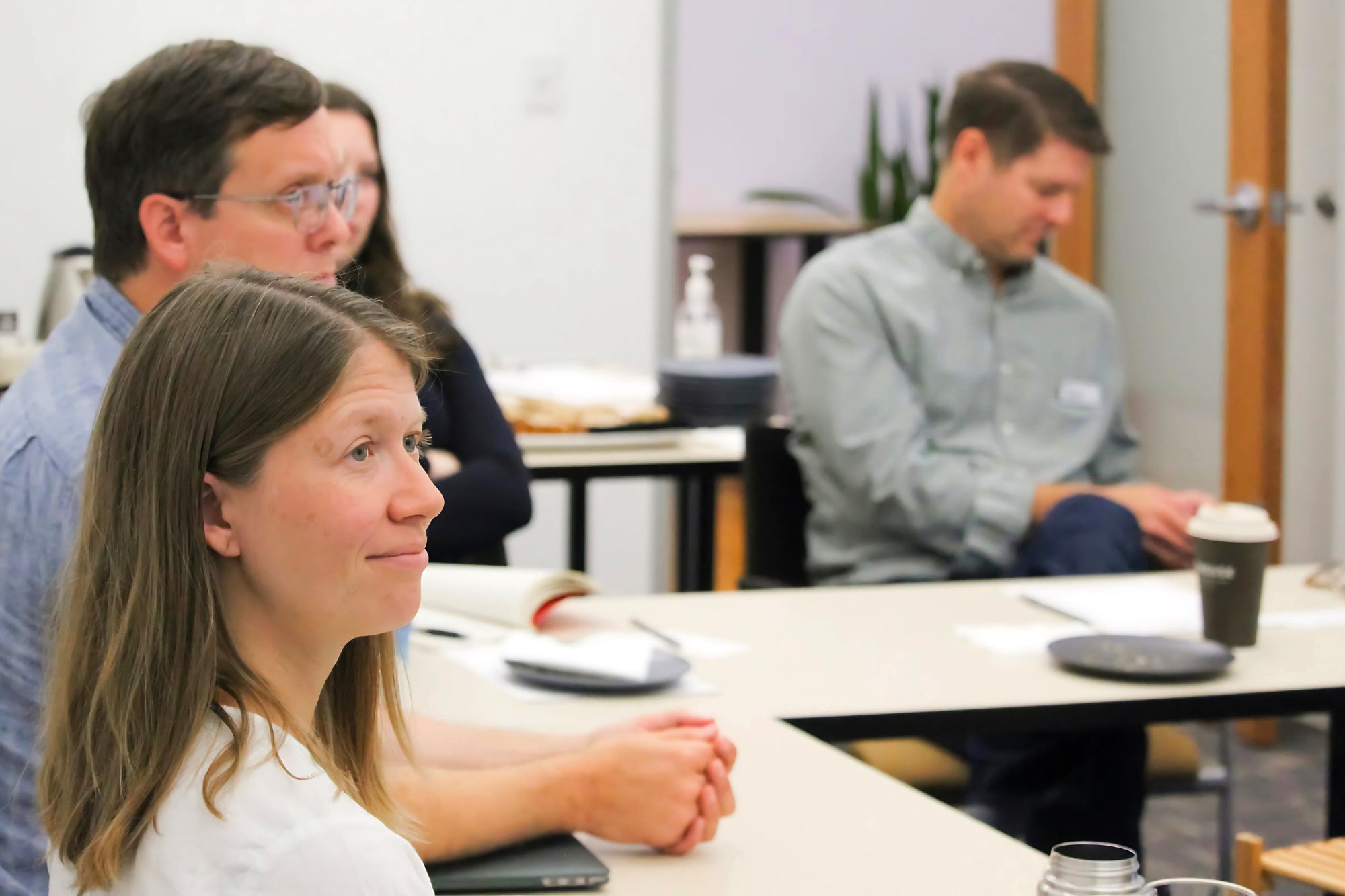 A group of people sit attentively in a meeting room, with a woman in the foreground listening closely and others focused on the discussion.