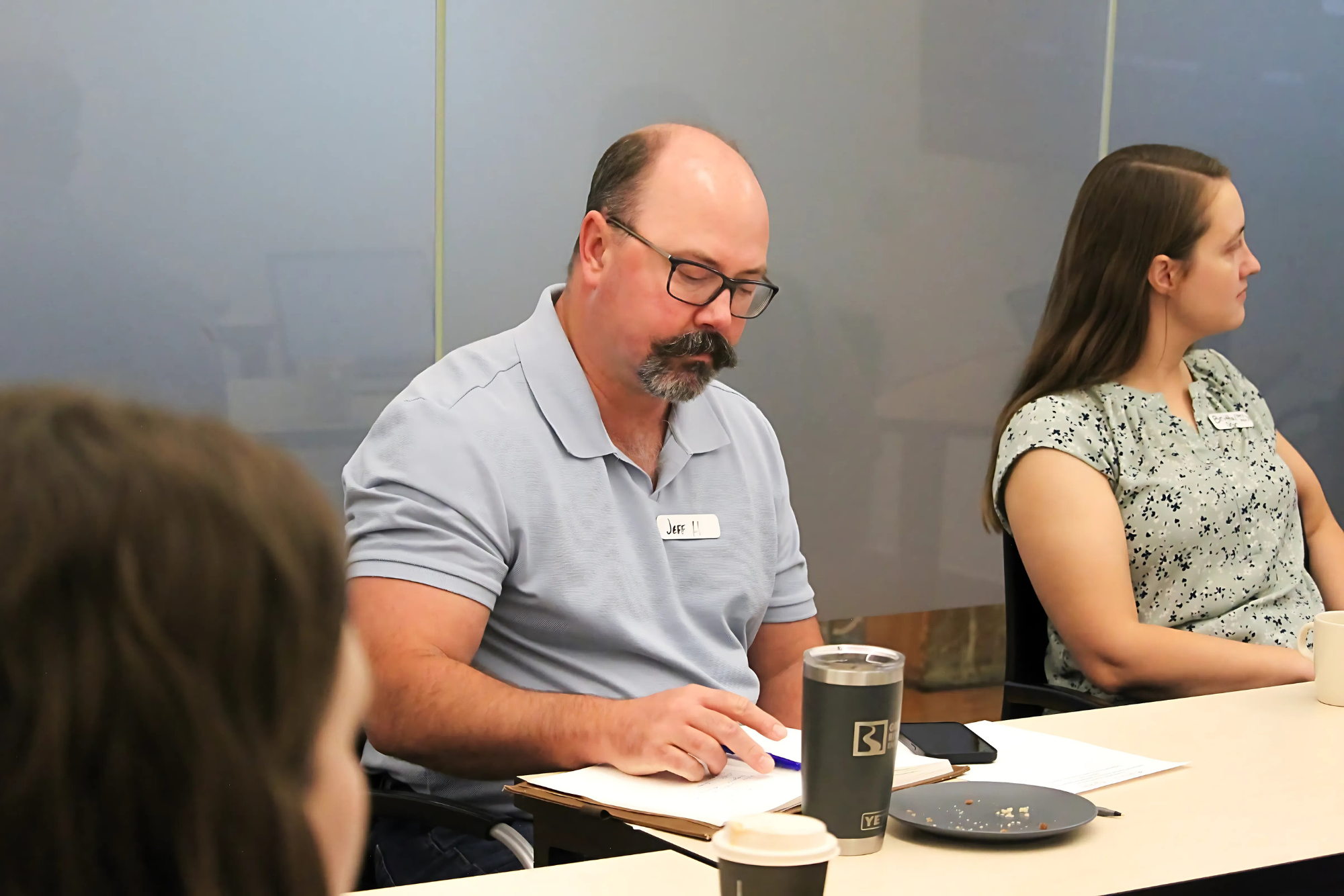 A man wearing a light blue shirt takes notes at a meeting table while seated next to a woman who is listening attentively.