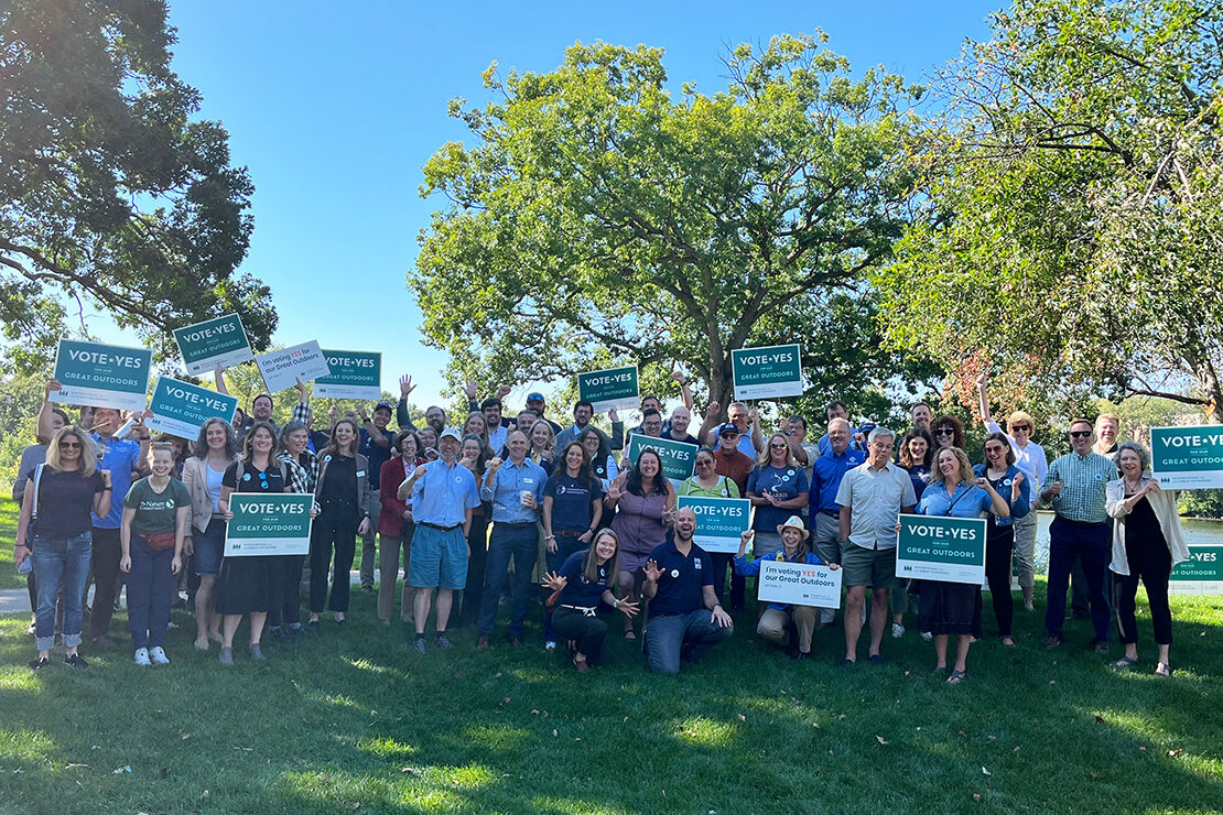 A large group of people standing on a grassy lawn outdoors on a sunny day, holding signs that read 'Vote Yes Great Outdoors.' The group appears cheerful, with many individuals smiling and waving at the camera. The background features trees with green foliage under a clear blue sky, creating a vibrant and positive atmosphere.