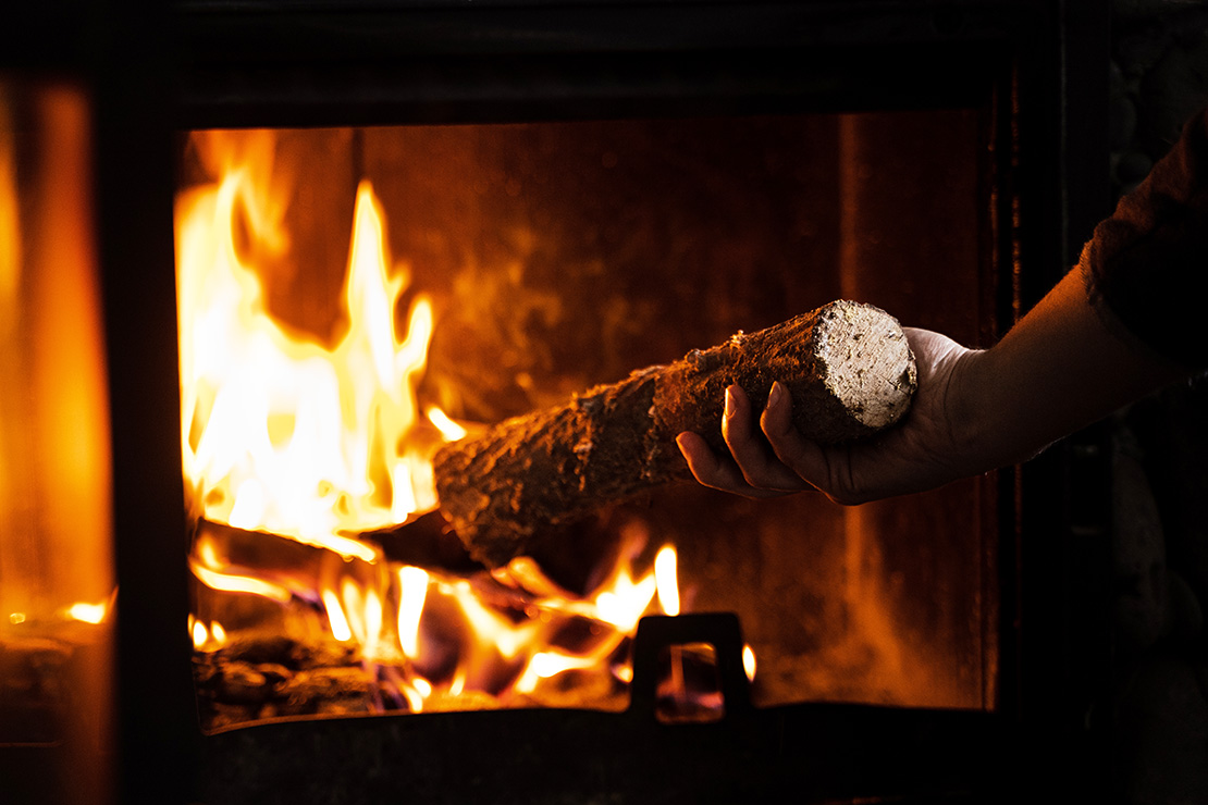 Hand placing a split log into a burning wood stove, with bright orange flames visible through the stove’s glass door.