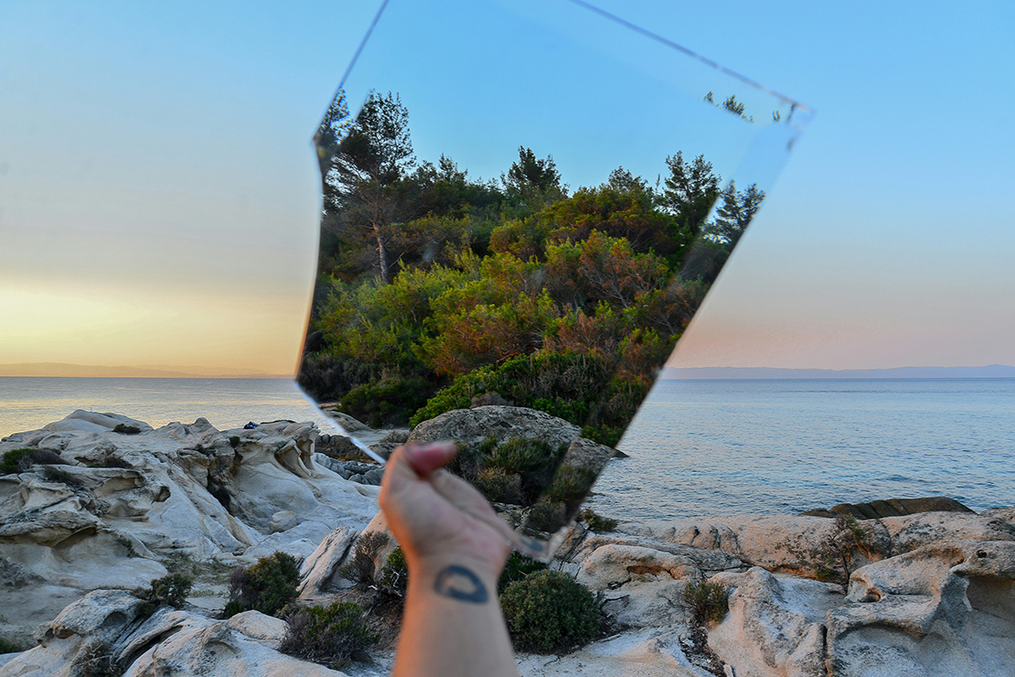 A hand holding a broken shard of mirror or glass in front of a rocky shoreline at sunset, the glass reflecting a cluster of green trees against a pastel sky while the actual background shows waves lapping the rocks under a clear blue horizon.
