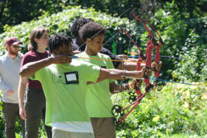 Young people in bright green shirts practice archery with compound bows in a sunlit, wooded meadow while other participants and staff watch from behind.