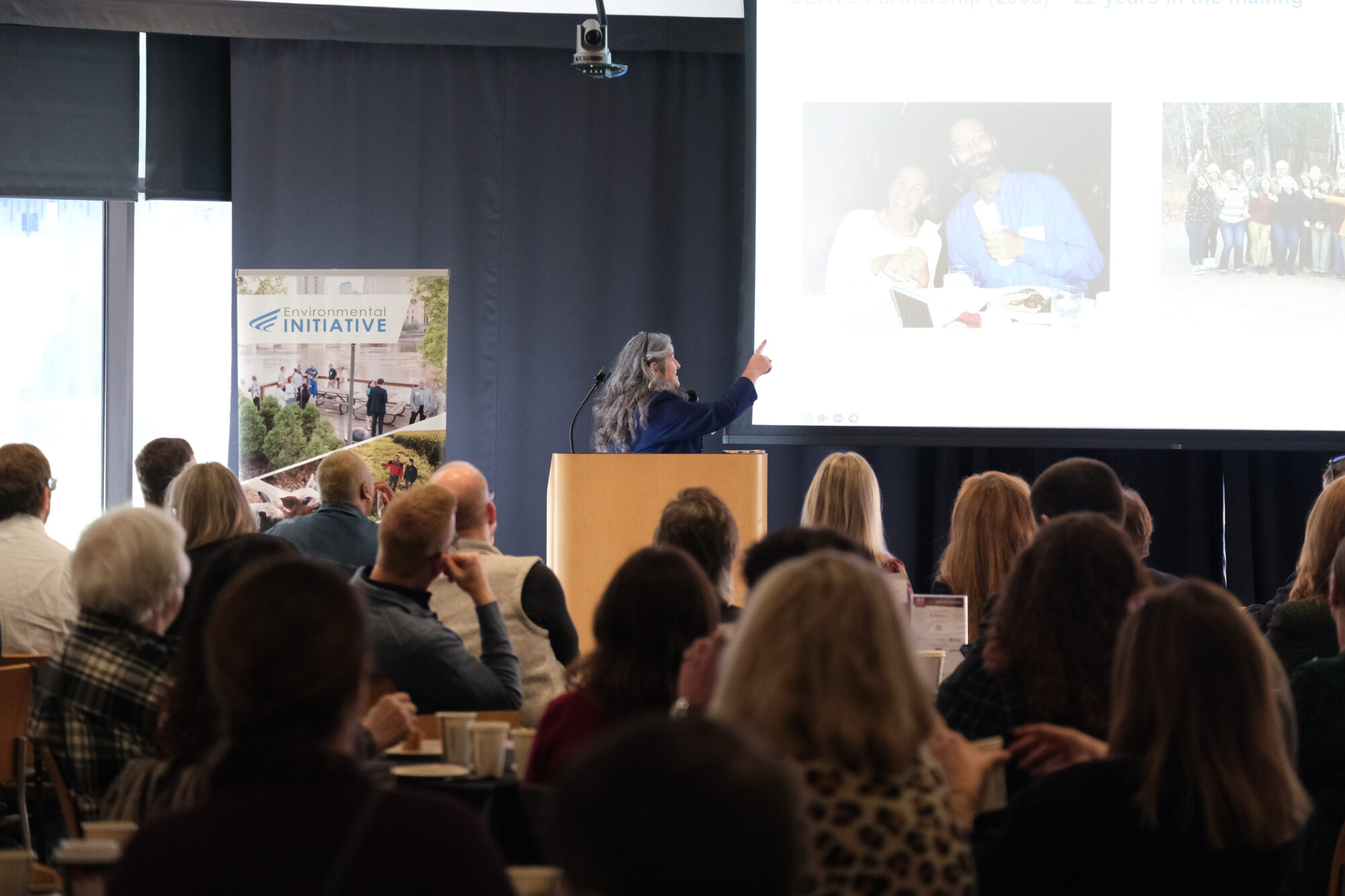 Speaker Diana McKeown of CERTs at the podium, pointing towards her slides projecting on the screen.