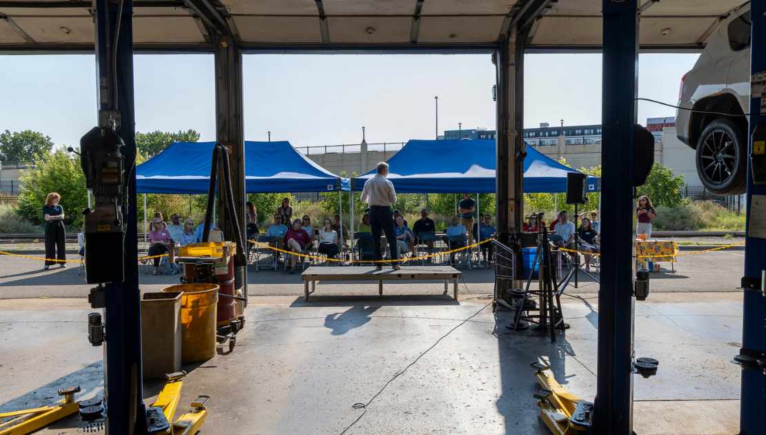 A man stands on a platform addressing a crowd in front of a garage bay.